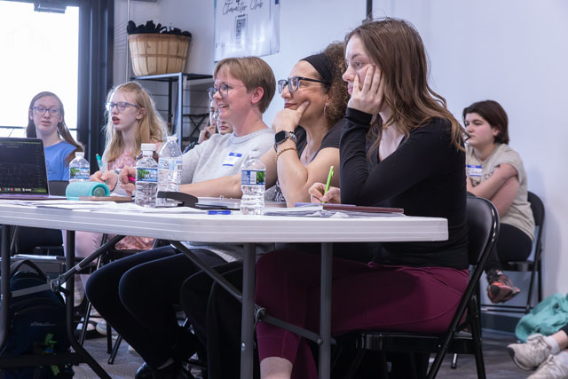 A group of people sitting at a table in a classroom setting, attentively listening to a presentation. Several participants have laptops and notebooks in front of them.