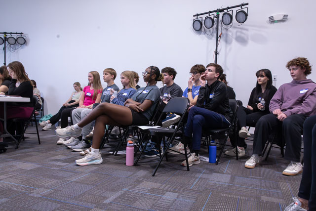 A group of young people sitting in classroom chairs, attentively listening during an event or meeting. Some are wearing name tags and holding water bottles.