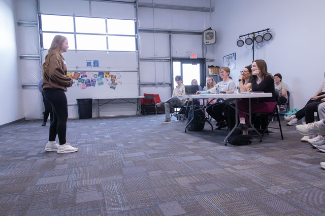 A young woman stands in front of a group of seated individuals in a classroom setting, delivering a presentation.