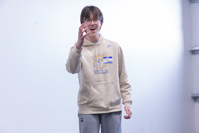 A smiling young man wearing a beige sweater with 'UCLA' printed on it and gray sweatpants, enthusiastically gesturing with one hand raised.