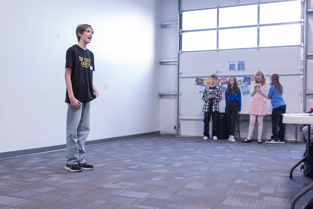 A young boy performs on a stage while several peers watch and react from behind him in a well-lit room.