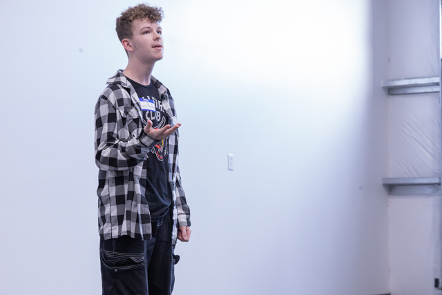 A young man with curly hair stands in front of a simple, white background, wearing a black and white checkered shirt over a black t-shirt. He appears to be speaking or performing, gesturing with one hand.