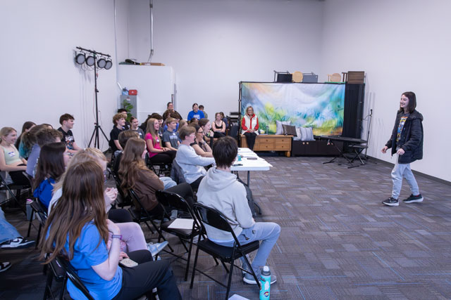 A group of young people seated in chairs watching a presenter at a workshop or class, with a backdrop of a colorful scene visible behind the speaker.