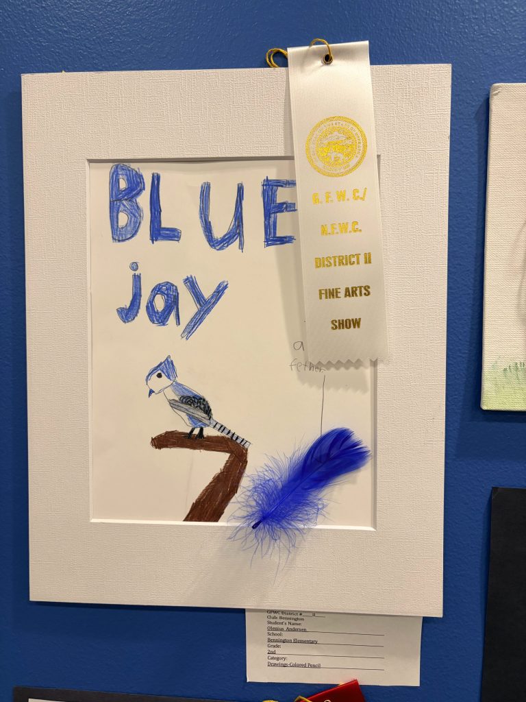 A framed artwork featuring a drawing of a blue jay on a branch, with the title 'BLUE jay' in blue letters. The artwork is decorated with a blue feather and includes a ribbon identifying it as an award from a fine arts show.