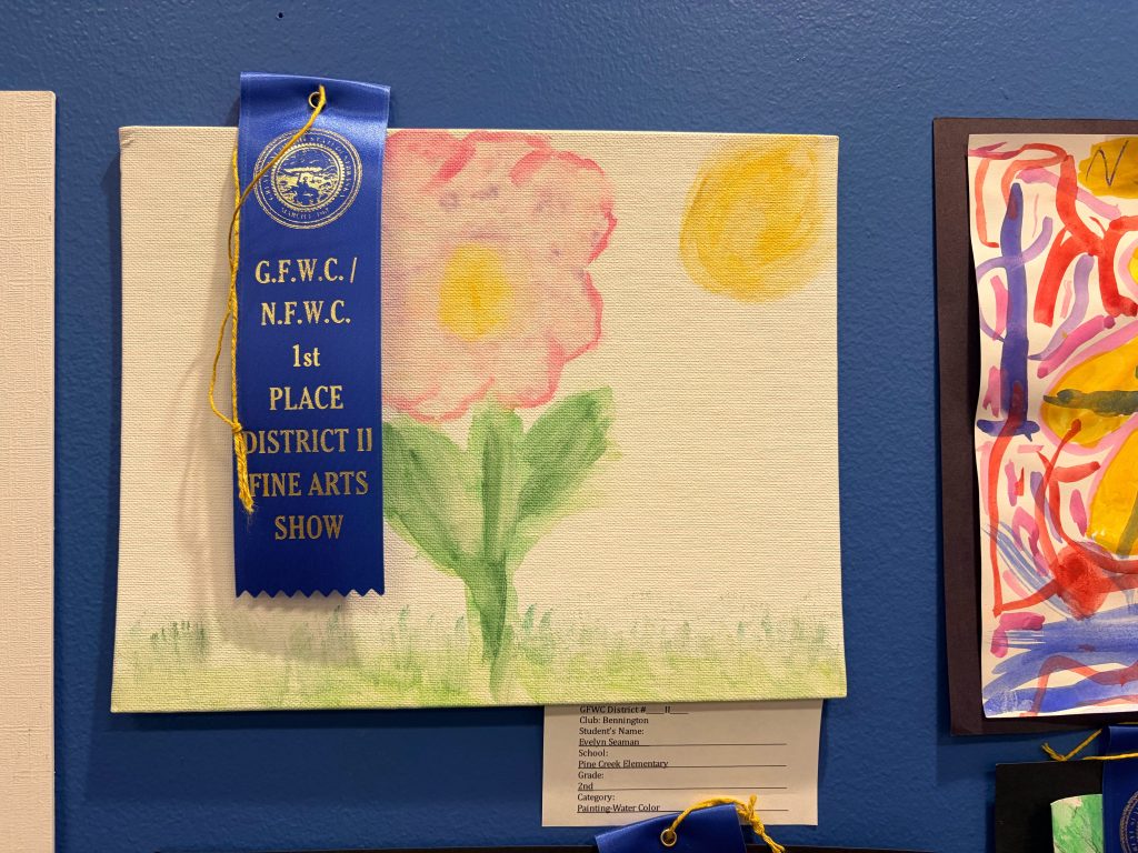 A first place ribbon awarded for a flower painting displayed at a fine arts show, featuring a pink flower and a yellow sun on a canvas.