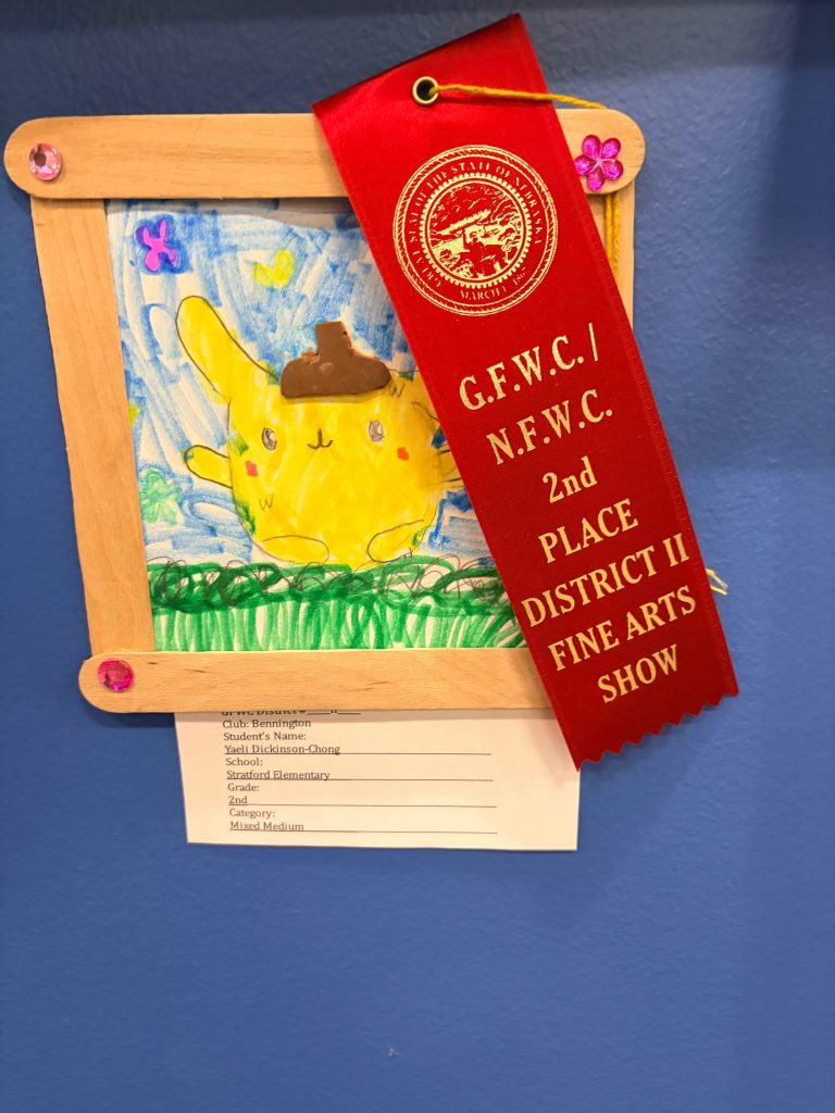 A framed drawing of a yellow cartoon character with a brown hat, surrounded by colorful flowers and grass, displayed with a red ribbon that reads '2nd PLACE DISTRICT II FINE ARTS SHOW'.