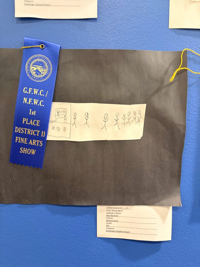 Artwork featuring a drawing by a student, displaying stick figures and a small booth, with a blue ribbon indicating 1st place in a district fine arts show.