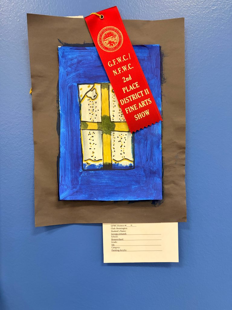 A colorful artwork featuring a window with a yellow cross and decorative details, displayed on a brown paper background. The piece has a red ribbon labeled '2nd PLACE DISTRICT II FINE ARTS SHOW' attached to it.