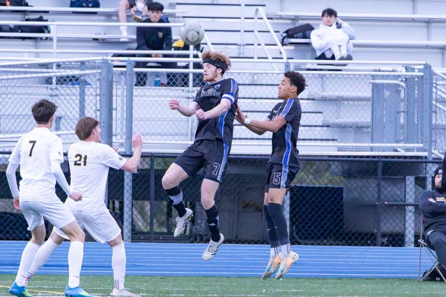 Two soccer players in black jerseys jumping to head a ball, while two players in white jerseys watch from the ground.