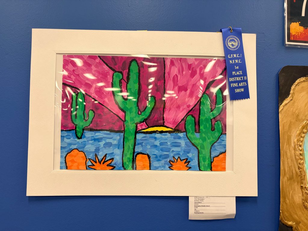 A colorful painting depicting cacti and a sunset over a body of water, framed and displayed with a blue ribbon for first place in a fine arts show.