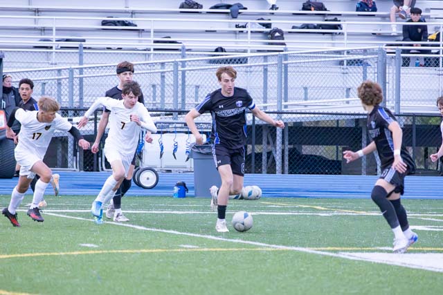 A group of young male soccer players on a field, with one player in a black uniform dribbling a soccer ball, while others in white chase after him.