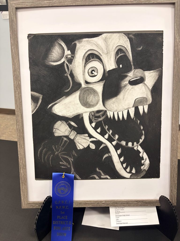 A charcoal drawing of a cartoonish, monstrous character with exaggerated features, showing sharp teeth and wide eyes. The artwork is framed and accompanied by a blue ribbon designating it as '1st Place' in a district fine arts show.
