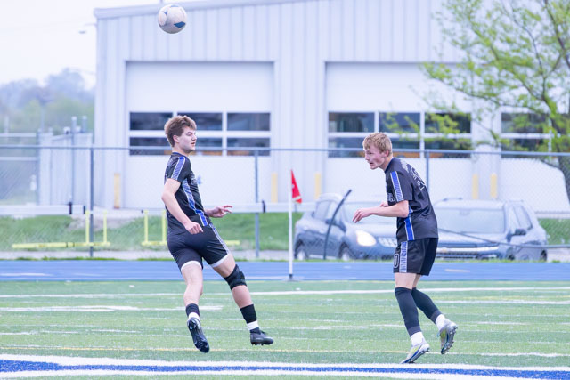 Two male soccer players in black uniforms, one about to kick a soccer ball in the air while the other is ready to receive it, on a green turf field.