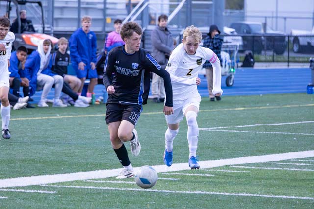 Two high school soccer players compete for the ball on a grassy field, with teammates and spectators in the background.