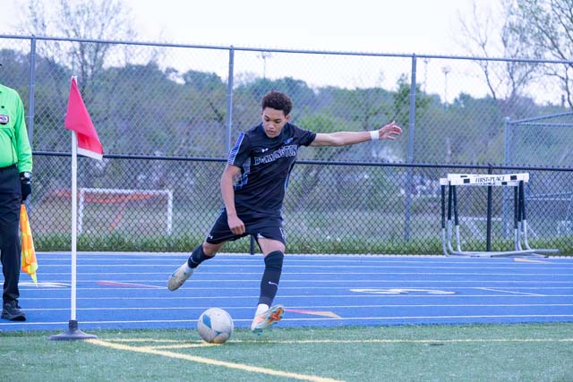 A young soccer player in a black uniform prepares to take a corner kick during a match, with a referee and field equipment visible in the background.