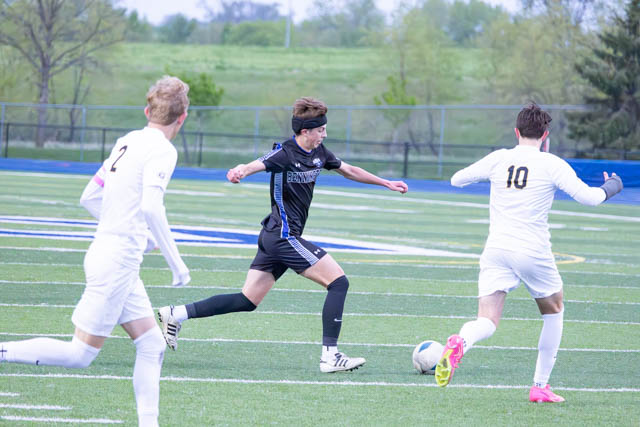 A soccer player in a black jersey dribbles the ball on a field, pursued by two opponents wearing white jerseys. The background shows a grassy area and trees.