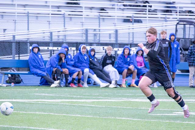 A soccer player in a black uniform with the number 25 is sprinting on the field towards a soccer ball, while several spectators in casual attire observe from the sidelines under overcast weather.