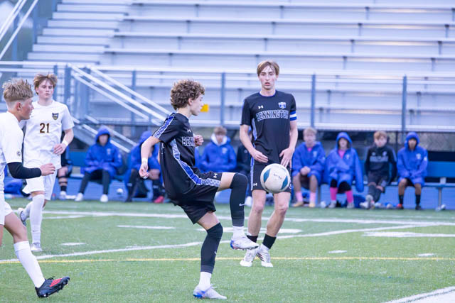 A youth soccer match in progress with players from different teams. A player in a black jersey is about to kick the ball, while another player in a white jersey and a few spectators can be seen in the background.