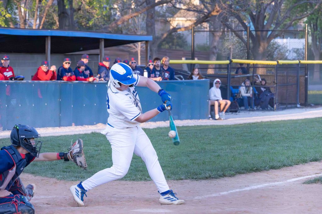 A baseball player in a white uniform swings a bat at a pitch, with a catcher in a black helmet and chest protector ready to catch. Fans are visible in the background.