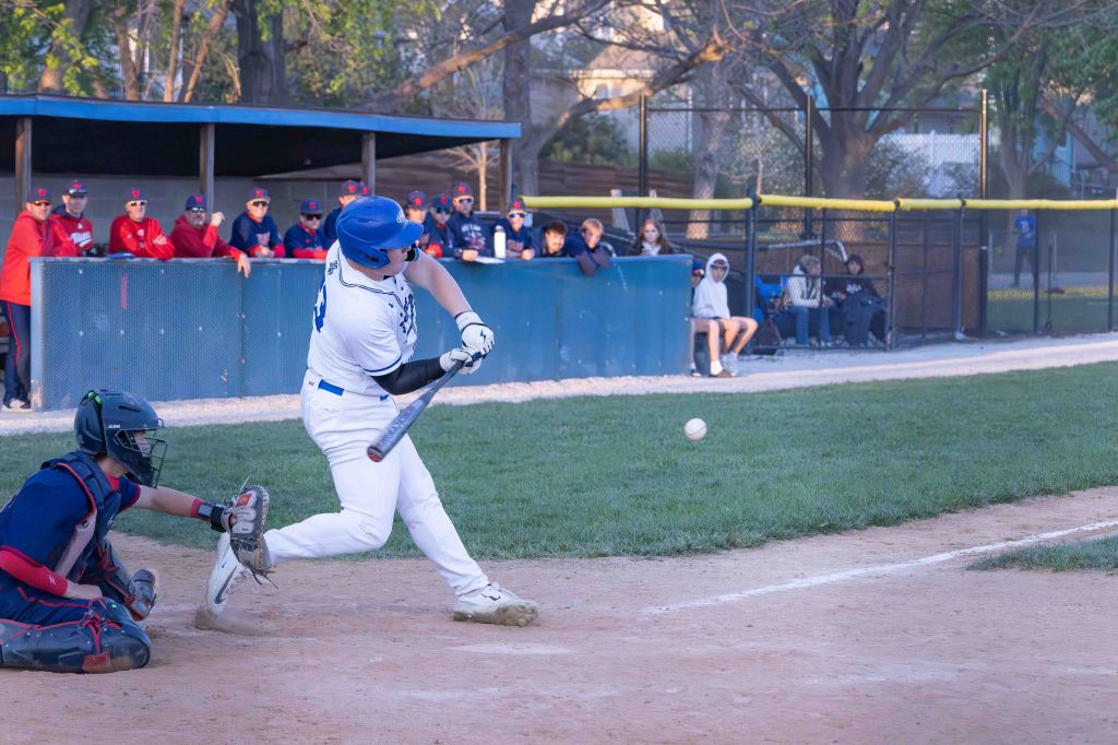 A baseball player in a white uniform swings at a pitch while a catcher crouches behind home plate. A group of spectators can be seen in the background.