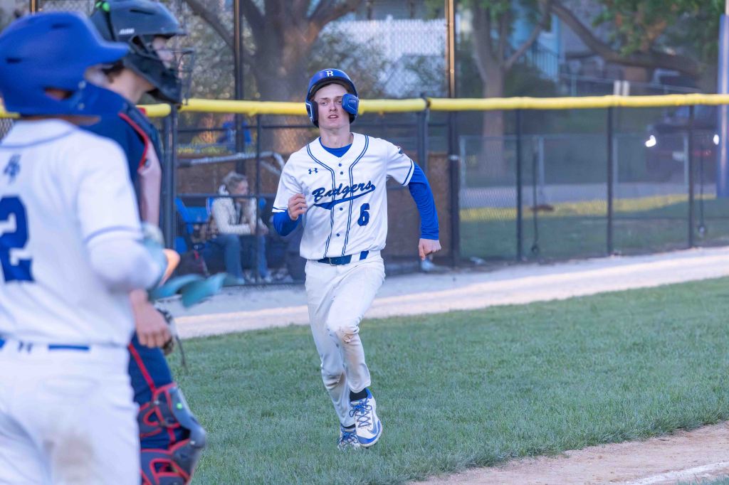 A baseball player in a white uniform with blue accents is running towards home plate, celebrating a run. The field is green with a yellow outfield fence in the background.
