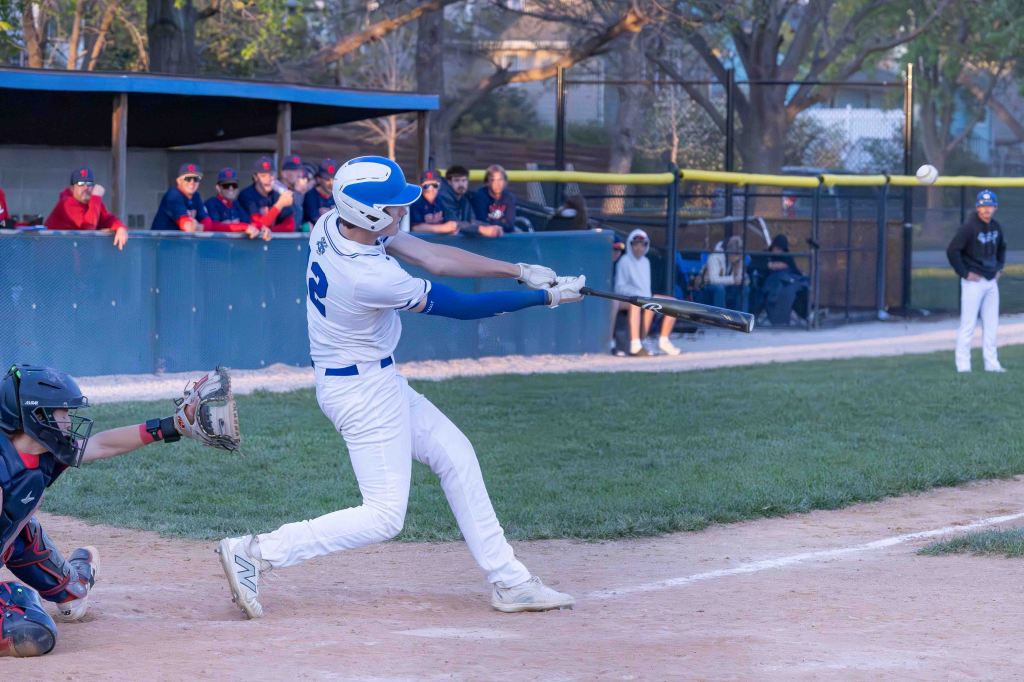 A baseball player in a white uniform swings a bat at a pitch, while a catcher in a black uniform crouches behind home plate. Spectators in the background watch the game.