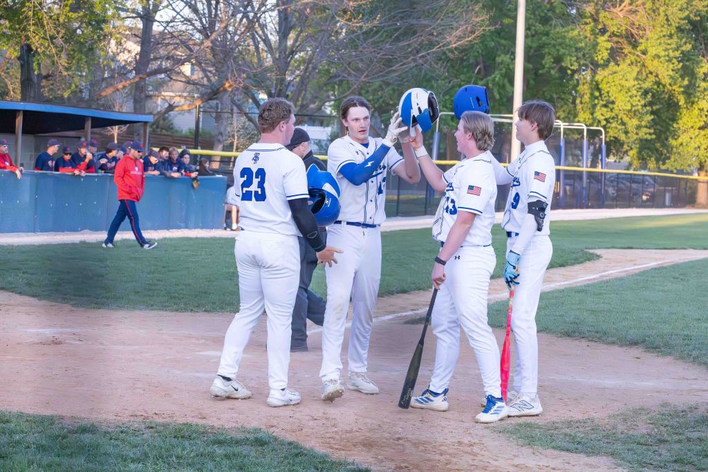 A group of four baseball players in white uniforms celebrate at home plate. They are exchanging helmets and discussing plays, with a cheering crowd visible in the background.