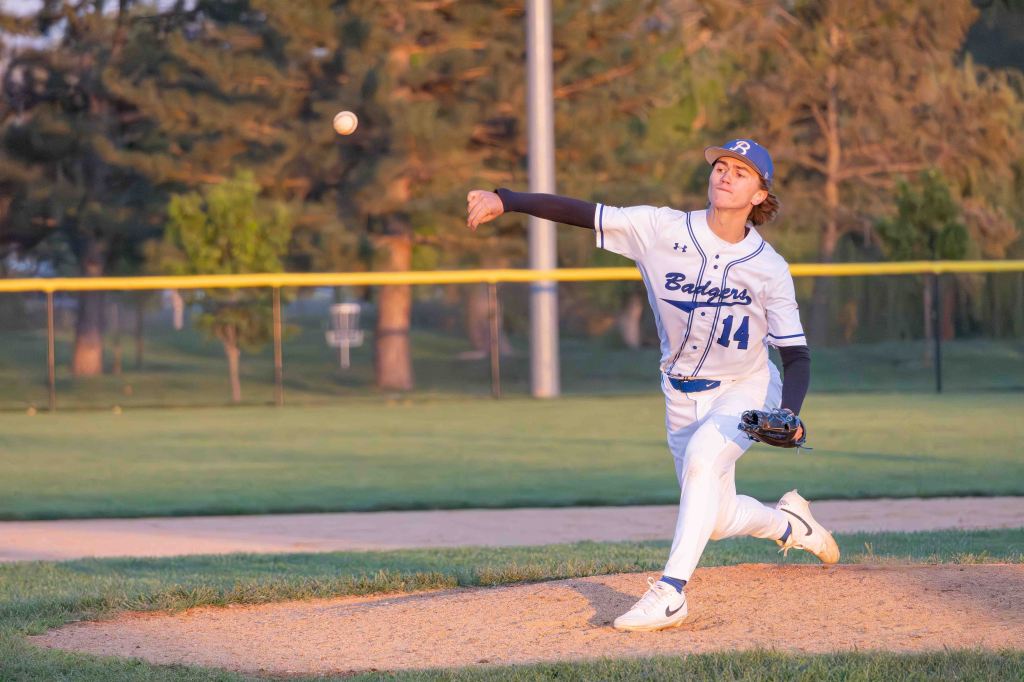 A young baseball pitcher in a white and blue uniform throws a pitch on a baseball field during sunset.