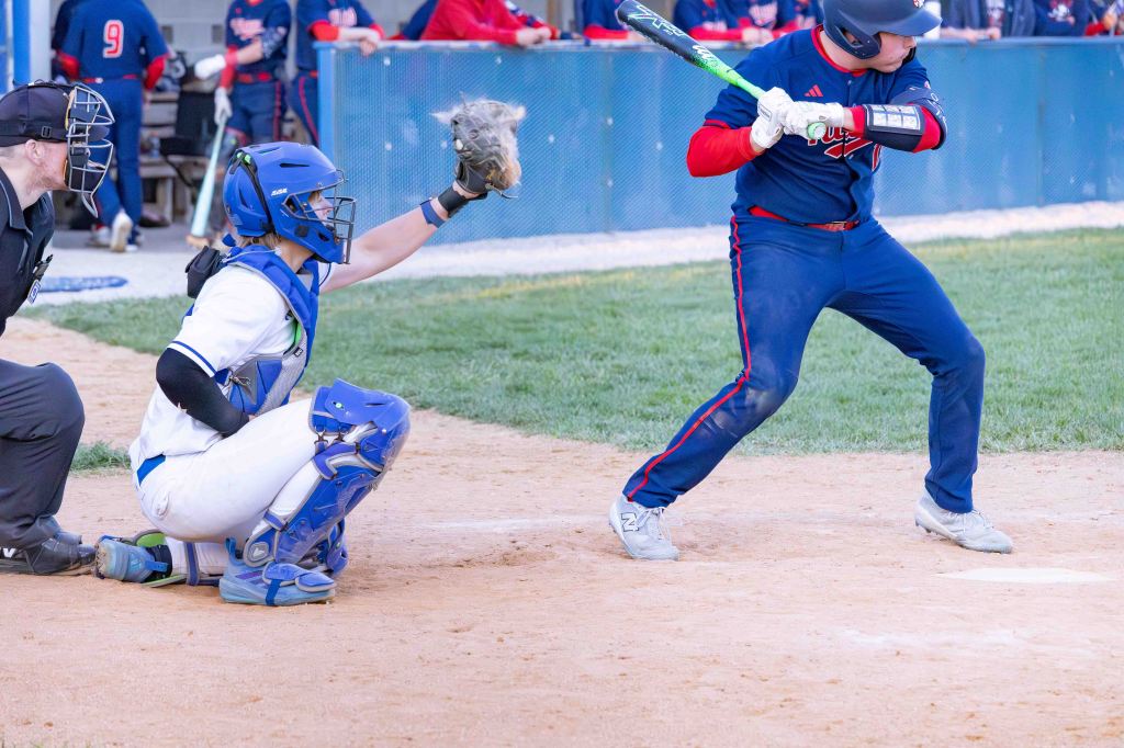 A baseball player prepares to bat while a catcher in blue gear crouches behind home plate. An umpire stands nearby, observing the play.