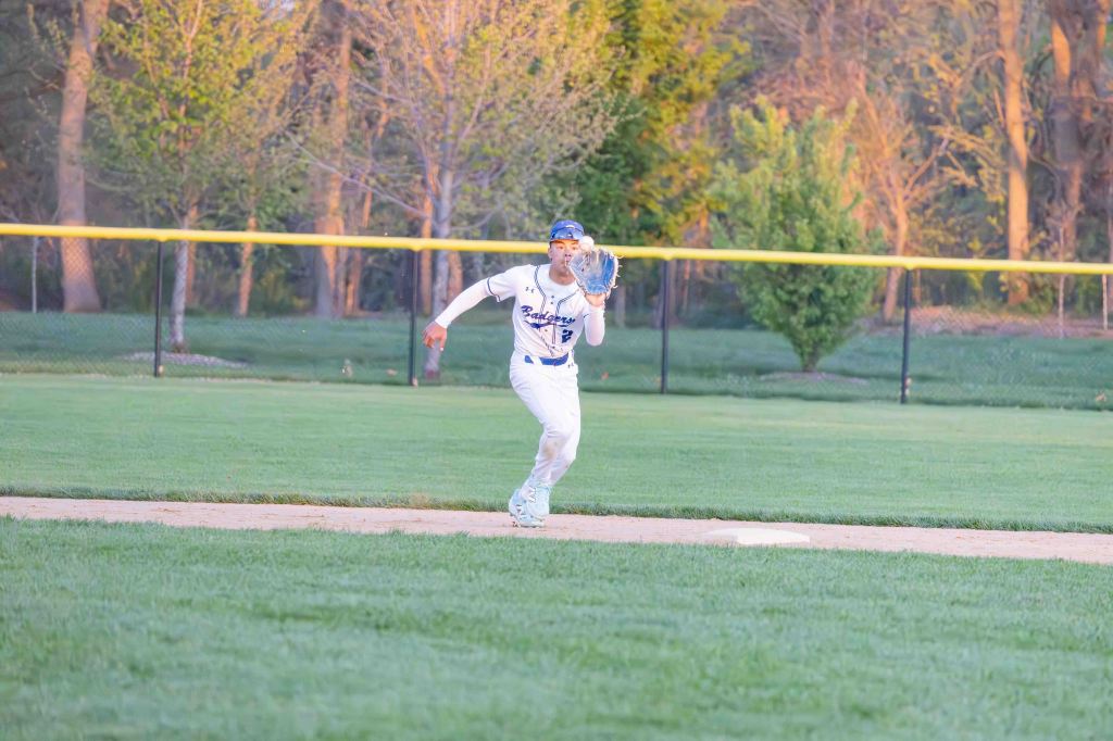 A baseball player in a white uniform prepares to catch a ball in an outdoor field during a game, with a batting area and fence visible in the background.