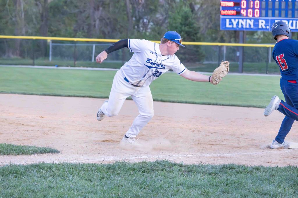 A baseball player in a white uniform reaches for the ball while attempting to tag a runner on a dirt infield during a game.