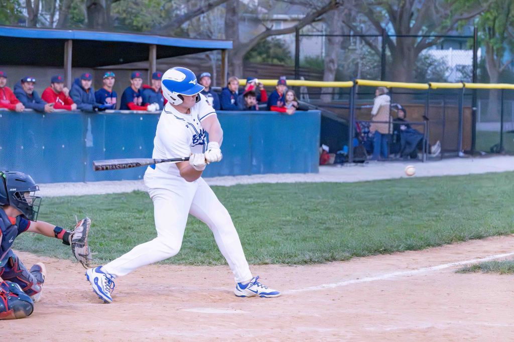 A baseball player in a white uniform swings a bat to hit a ball while a catcher crouches behind home plate, with spectators visible in the background.