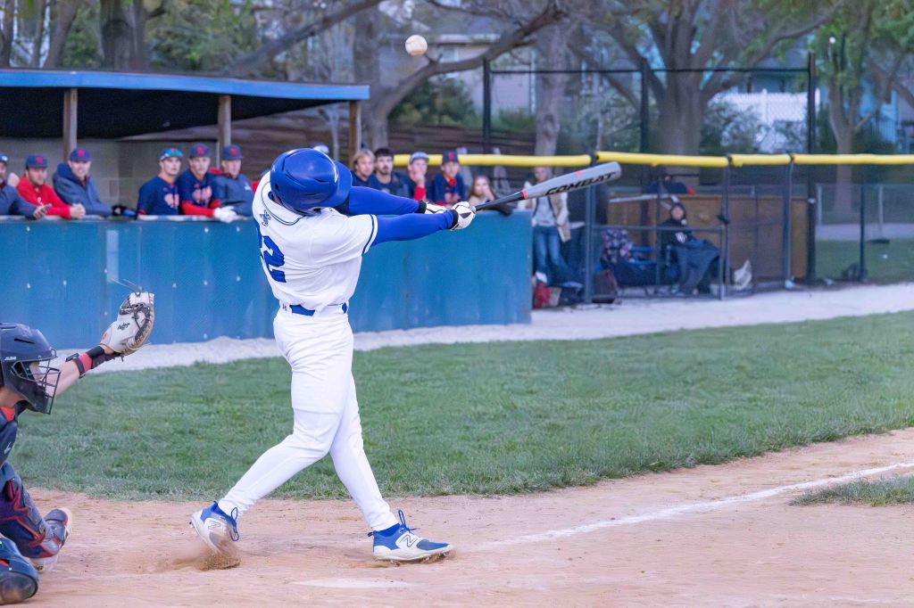 A baseball player in a white uniform swings a bat at a flying ball during a game, with spectators in the background.