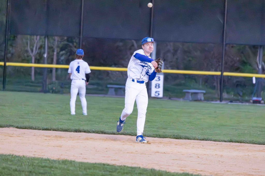 A baseball player in a blue and white uniform is throwing a baseball while standing on the infield, with another player in the background.