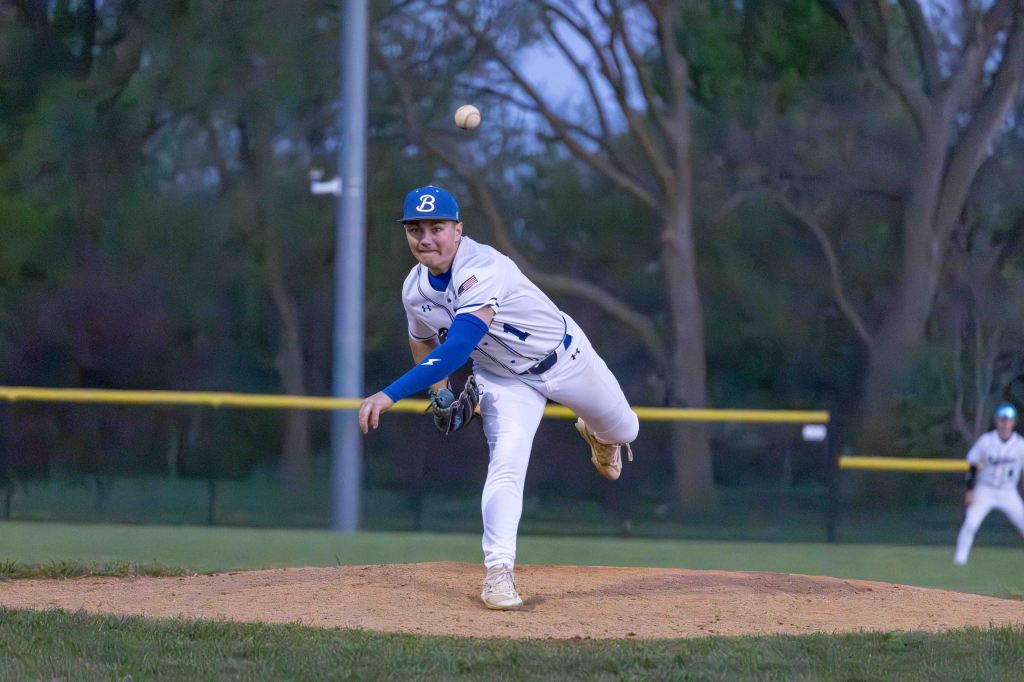 A young baseball pitcher in a white uniform and blue cap is delivering a pitch from the mound during a game, with trees in the background and a teammate visible in the outfield.