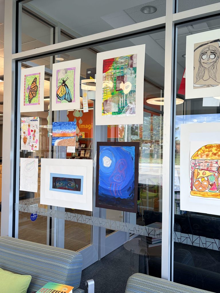 A display of children's artwork in a library window, featuring various colorful drawings and paintings, including a butterfly, abstract art, and a whimsical food illustration.