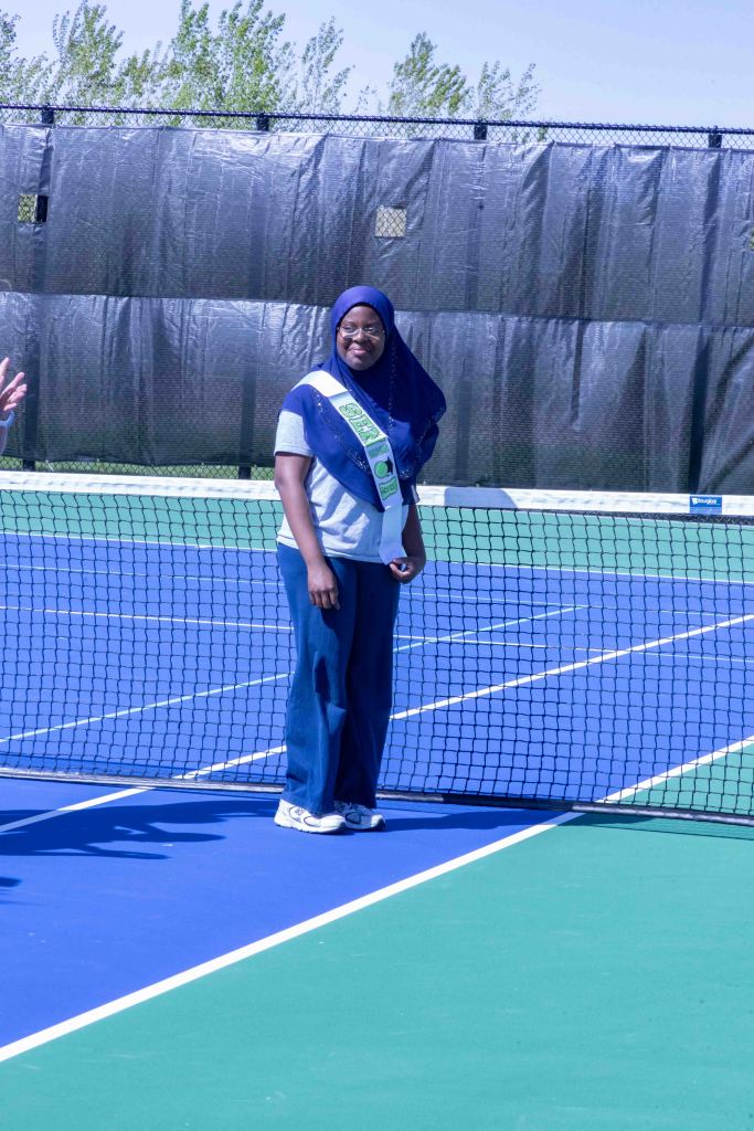 A person wearing a hijab and casual clothing stands on a tennis court near the net, smiling and looking towards the camera.