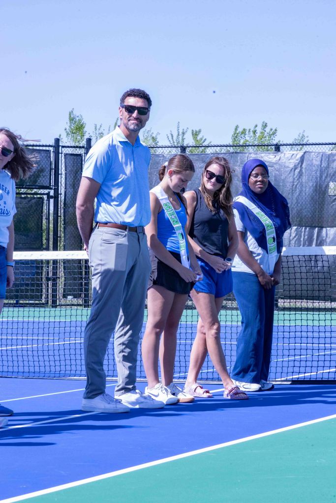 Group of individuals standing on a tennis court, including a man in a blue polo shirt and gray pants, and several women wearing casual attire and sashes, in bright sunlight with a net in the background.