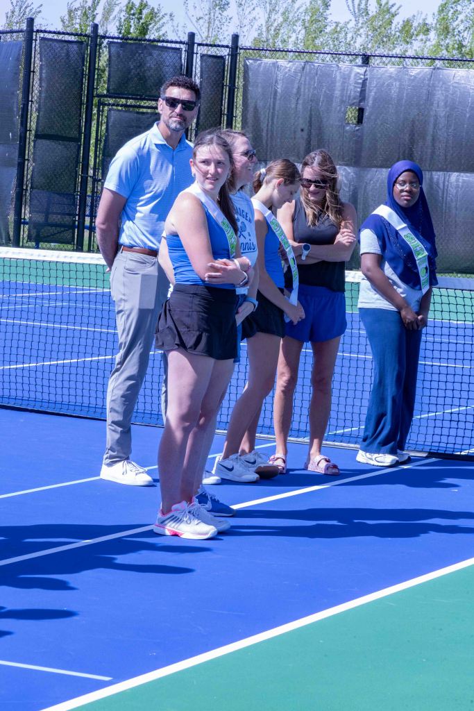 A group of tennis players and a coach standing on a tennis court, wearing sports attire. They are positioned near the net with a clear blue sky in the background.