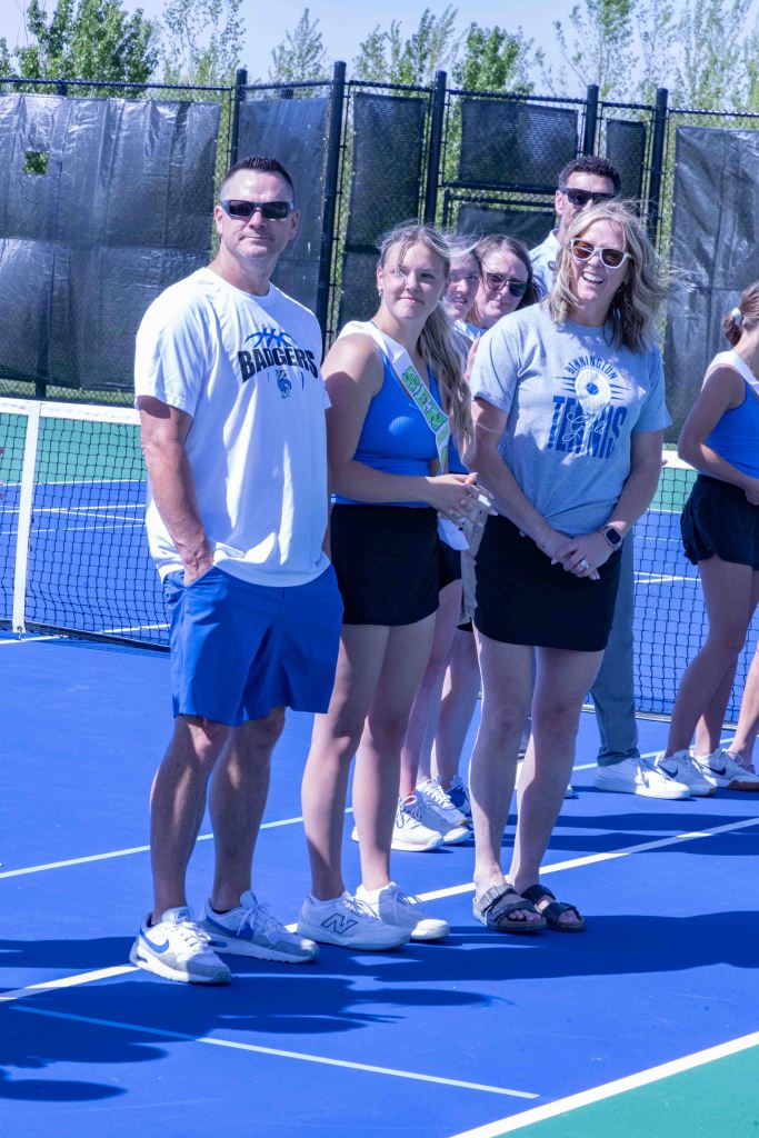 A group of people standing on a blue tennis court, including a man in a white shirt and sunglasses, a young woman in a blue top with a sash, and another woman in a gray shirt. The background features tennis nets and green trees.
