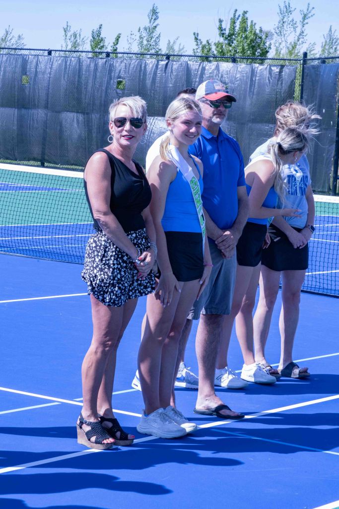 A group of people standing in line on a tennis court, with a focus on a young girl wearing a sash and a woman in sunglasses and a black top with a floral skirt.