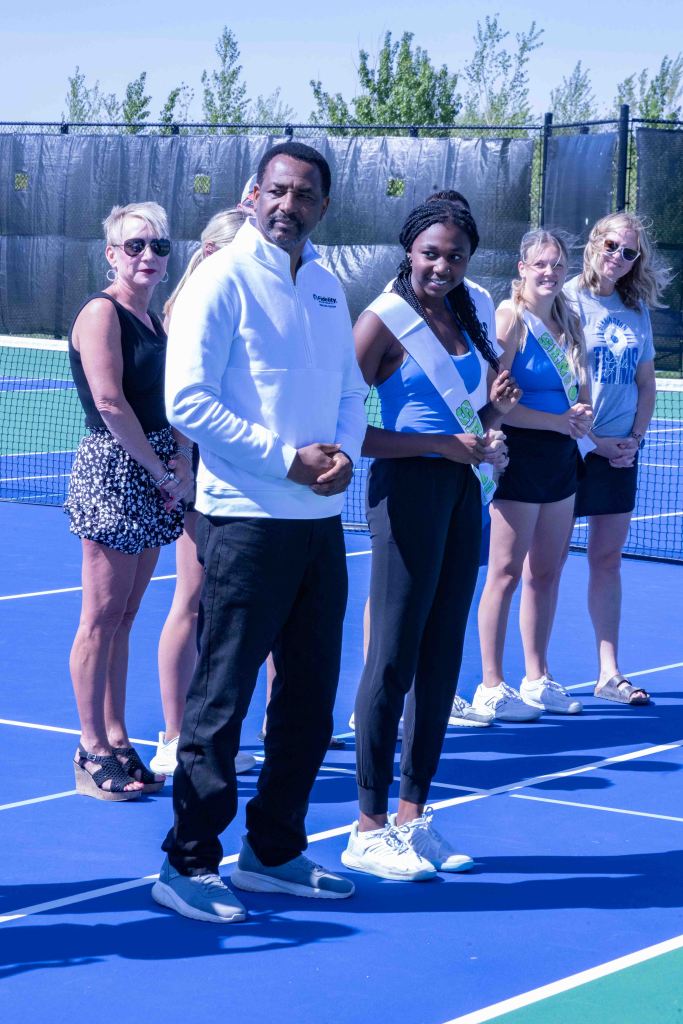 A group of people standing on a tennis court, including a man in a white jacket, a woman in a blue top wearing a sash, and others in athletic clothing.