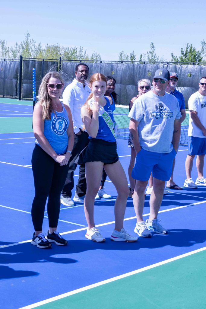 Group of people standing on a tennis court, smiling and interacting. Several individuals are wearing sports attire, and one is holding a sash. The background features tennis nets and greenery.