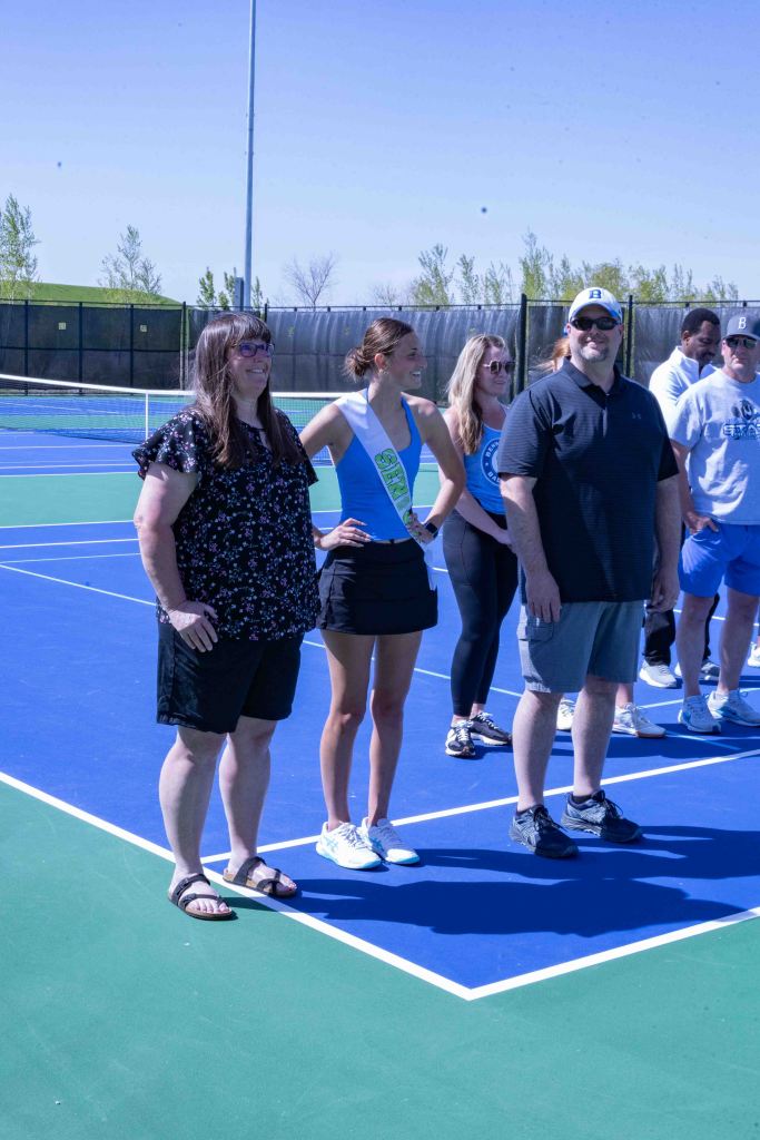 Group of people posing on a tennis court, including a woman wearing a sash, indicating a special title or position.