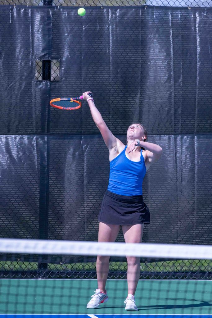 A young female tennis player serving the ball on a court, wearing a blue tank top and a black skirt.