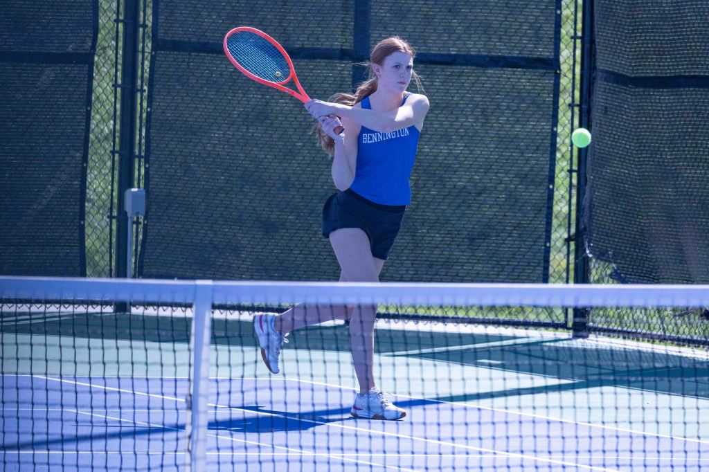 A female tennis player in a blue top is hitting a tennis ball on a court with a net and a fence in the background.