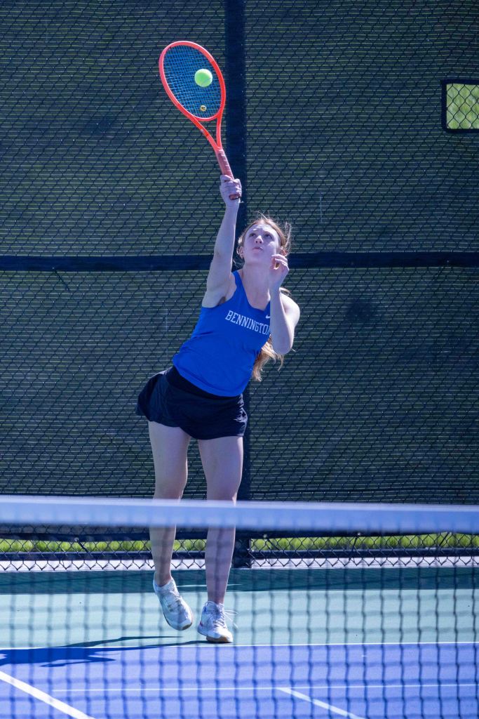A young woman serving a tennis ball on a court, dressed in a blue tank top and black skirt.