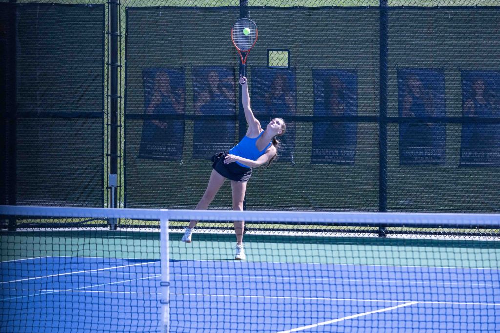 A female tennis player performing a serve on a court, with a green background and tennis net in view.