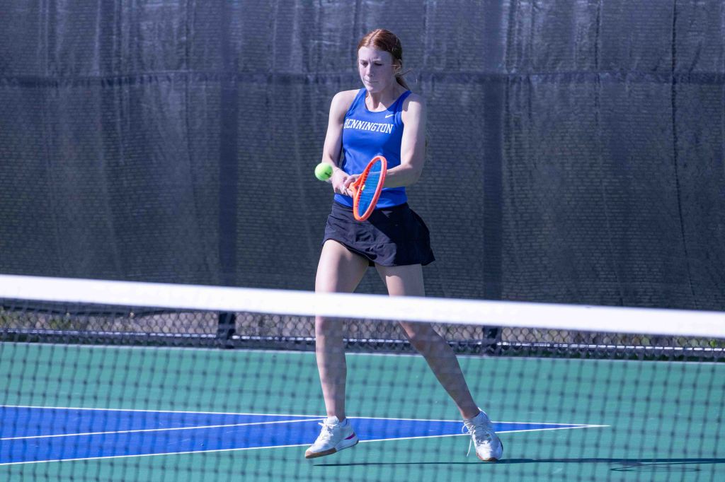 A female tennis player in a blue tank top and black skirt prepares to hit a tennis ball on an outdoor court.
