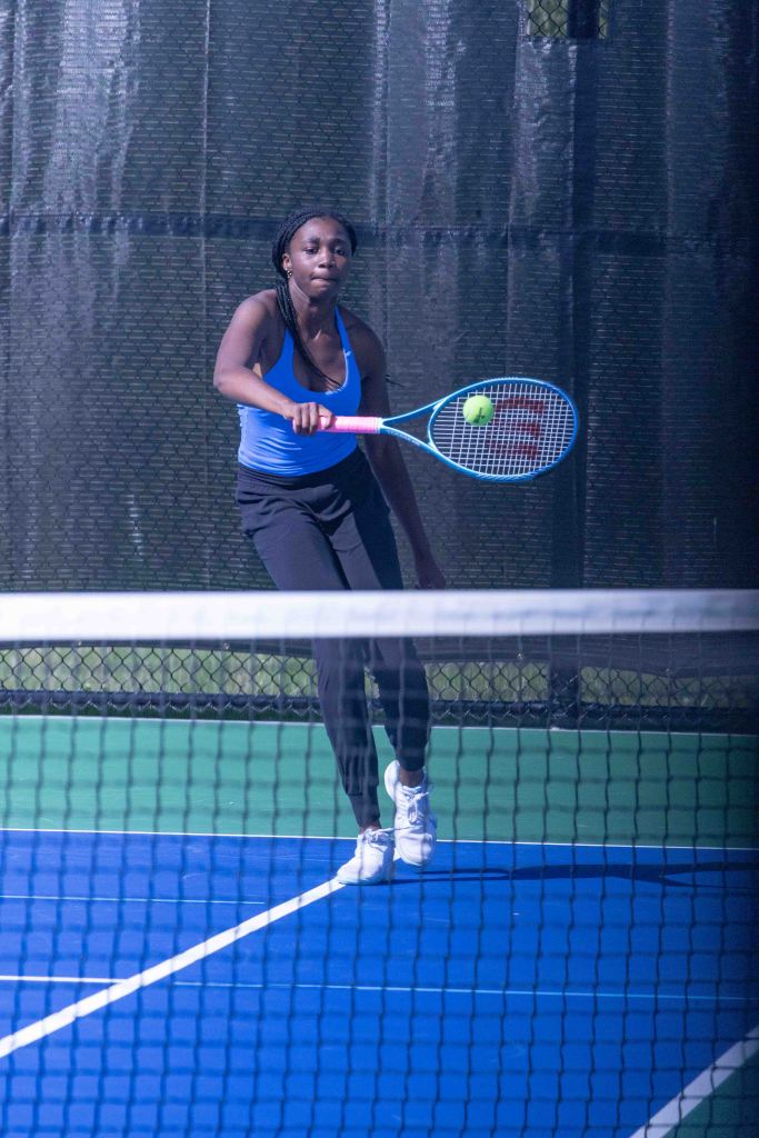 A young woman wearing a blue tank top and black leggings is poised to hit a tennis ball with a racket on a blue tennis court, with a net in the foreground.